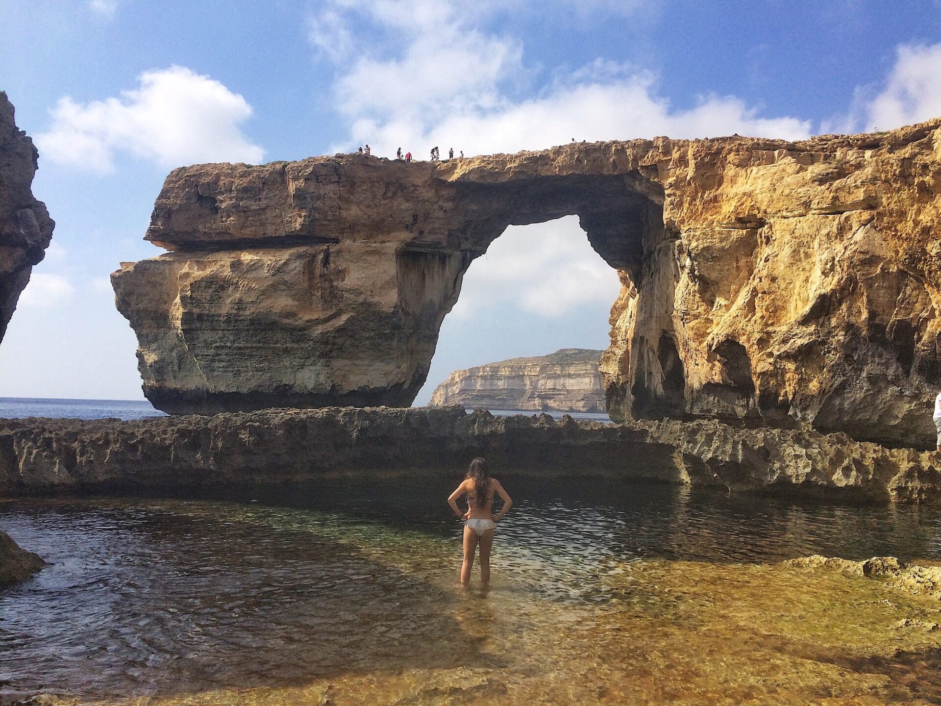 View of a girl standing in a tide pool looking in the distance at a large natural arch and cliffs over the ocean