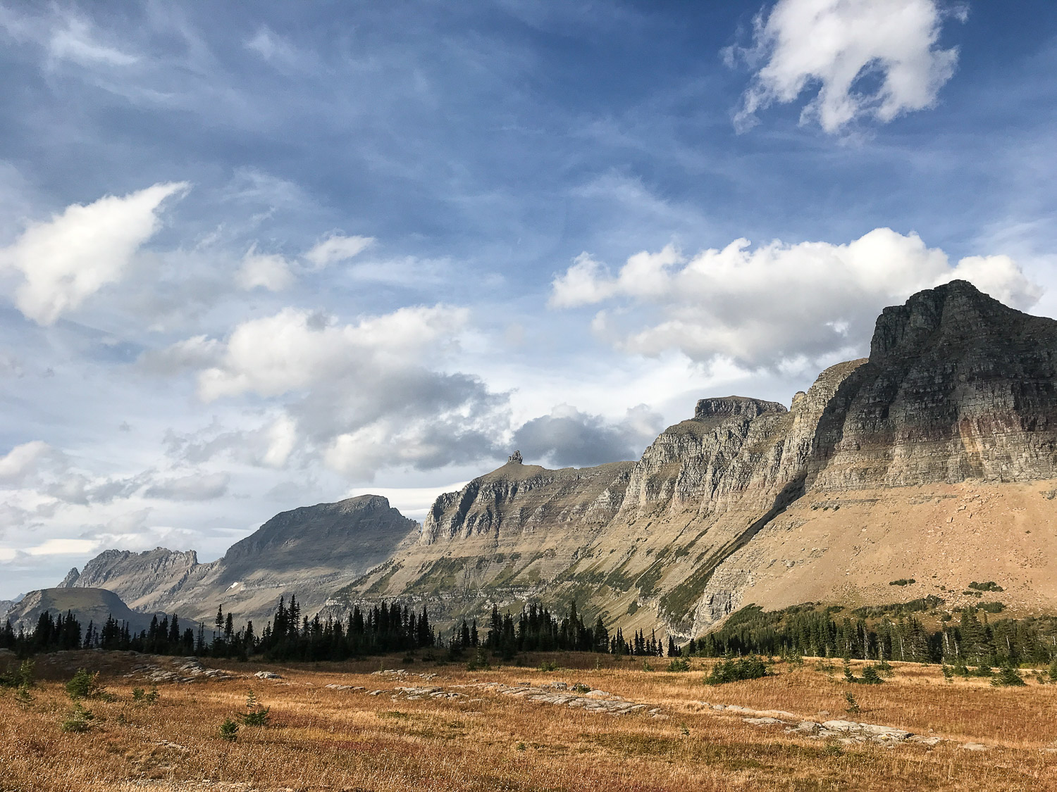 Blue skies and white clouds float over majestic mountains rising above the orange meadow in Glacier National Park