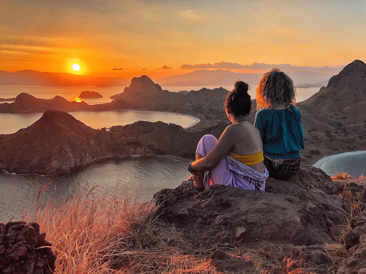 Two people sitting on the edge of a magnificent vista of islands and bays during the orange sunset at Komodo National Park