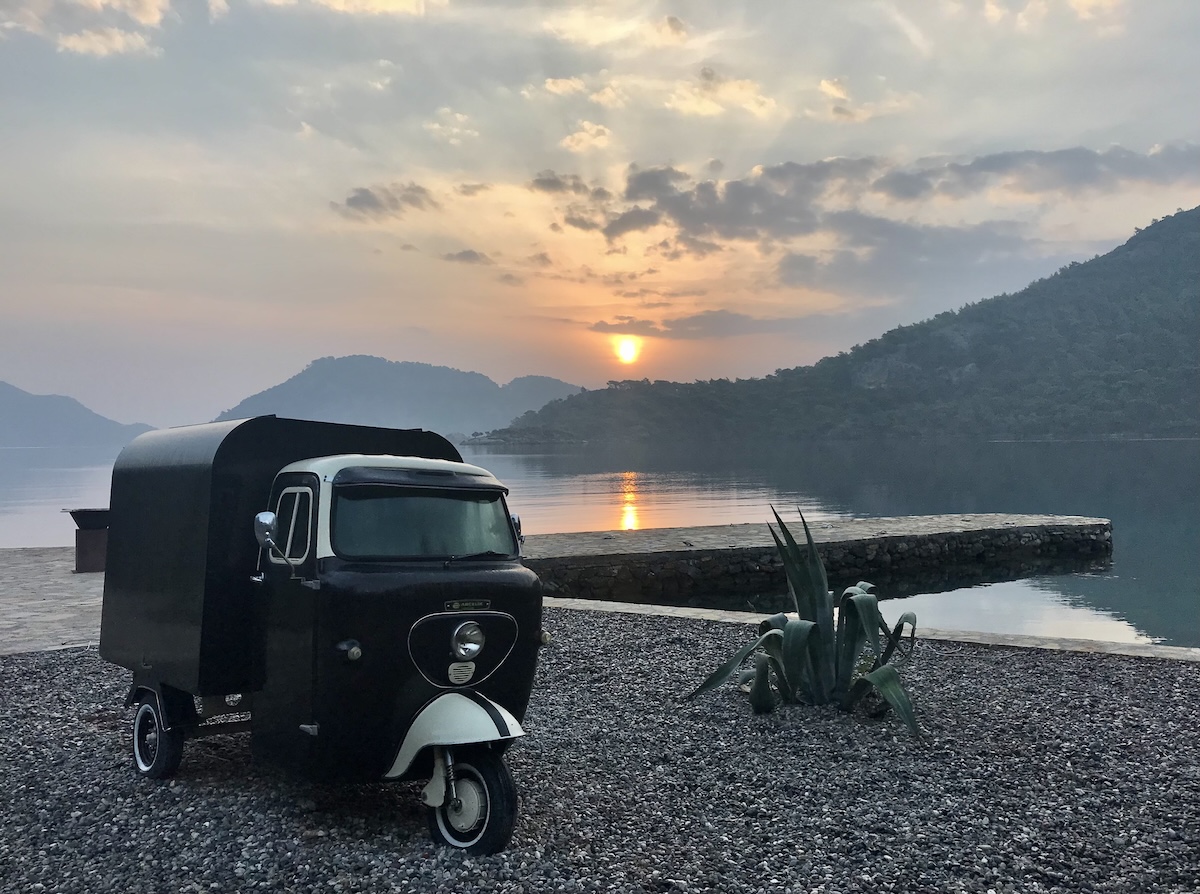 A tuk tuk sits an a gravel area on the sea with the sun rising over mountains in the background