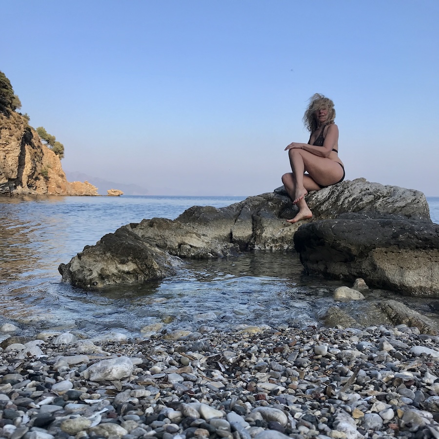 Woman sitting on rock in crystal clear Aegean sea by the pebbly shore
