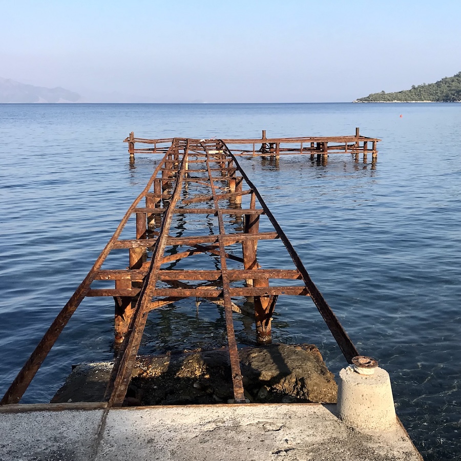 A weathered, rusty dock sits abandoned along the tranquil shores of Amos Bay, serving as a solitary relic amidst the untouched natural landscape.