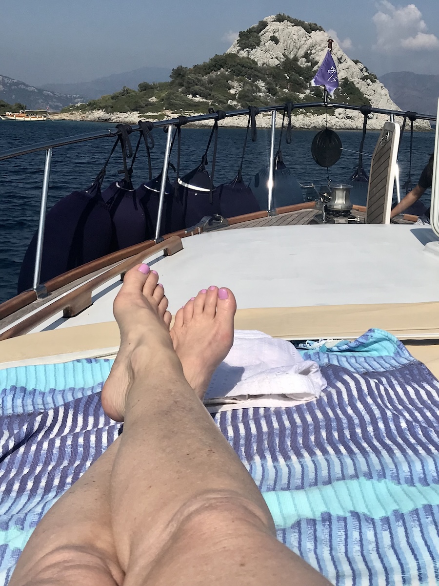 A young woman relaxing on the wooden deck of a boat, enjoying the serene view of the sparkling Aegean Sea under the bright Turkish sun.