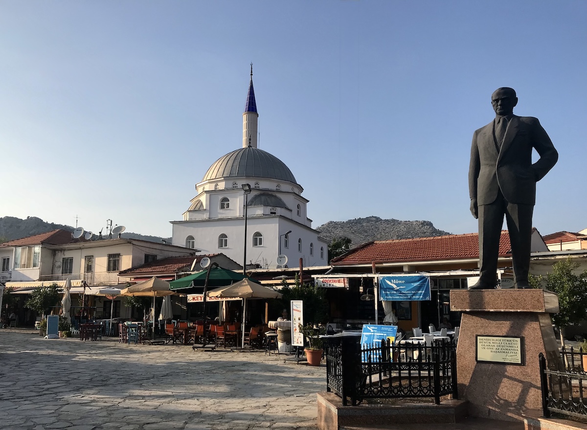 The quiet town square of Bozburun village, showing a stone statue of Mustafa Kemal Atatürk with a traditional mosque and minaret in the background