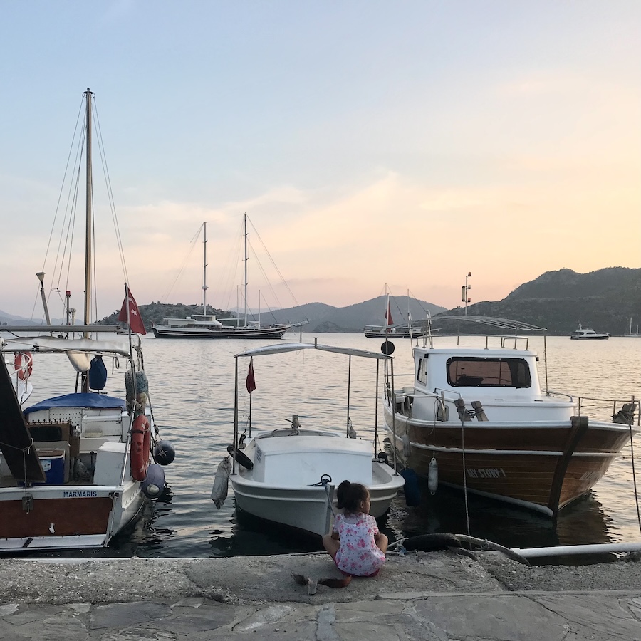 A young girl sits on a dock overlooking a calm harbor filled with boats during the golden hour of a quiet evening.